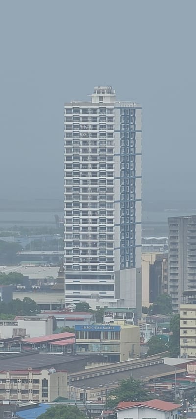 This is a high-angle, outdoor, eye-level shot of a cityscape in Makati, Philippines. The image is primarily focused on a large, rectangular building with a facade of numerous windows, suggesting it could be an office building or residential complex. The building is predominantly white and grey with blue accents. To the right of this main building, a long, elevated structure, possibly a walkway or an elevated road, runs parallel to the frame. This structure appears to have a grey, textured surface and is bordered by railings.

Further to the right, other buildings with varied architectural styles and colors are visible, including some with red roofs. There are also trees scattered throughout the scene, indicating a mix of urban development and greenery. A blue sign with white text that reads "MAGIC STAR SQUARE" is clearly visible on one of the buildings. The sky is overcast and hazy, suggesting it might be a cloudy day or that the photograph was taken during a time with lower visibility, perhaps morning or evening, or due to air pollution. No people are discernible in the image. The overall impression is that of a dense urban environment with multiple structures and the hint of a busy area.