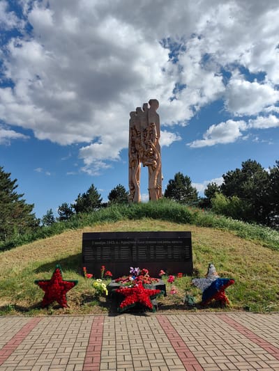 This is a photograph of a war memorial located on a grassy hill. The memorial consists of a tall, abstract sculpture of figures standing together, and a black plaque at the base of the hill with names and dates inscribed on it. In front of the plaque are two floral wreaths shaped like stars, one red and one in the colors of the Russian flag (white, blue, and red). Several red poppies are also visible scattered on the grass around the memorial. The sky above is partly cloudy with patches of blue. The foreground shows a paved walkway made of brick. The inscription on the plaque, in Russian, appears to be a list of residents who were burned alive on November 2, 1943, in a village called Kurotnoye. The overall impression is one of solemn remembrance.