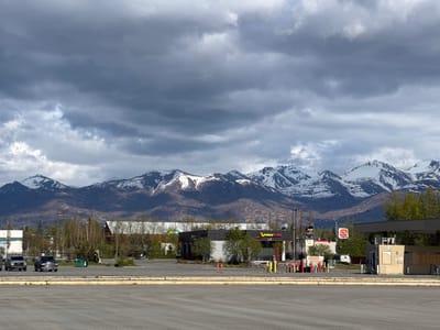 The image depicts a commercial area in Anchorage, Alaska, set against a backdrop of snow-capped mountains under a cloudy sky. In the foreground is a large, empty asphalt parking lot. Behind the parking lot, several commercial buildings are visible, including an Express Lube and a Burger King. Two pickup trucks are parked in the parking lot to the left. Utility poles and traffic lights are scattered throughout the scene. The sky is overcast with gray storm clouds.