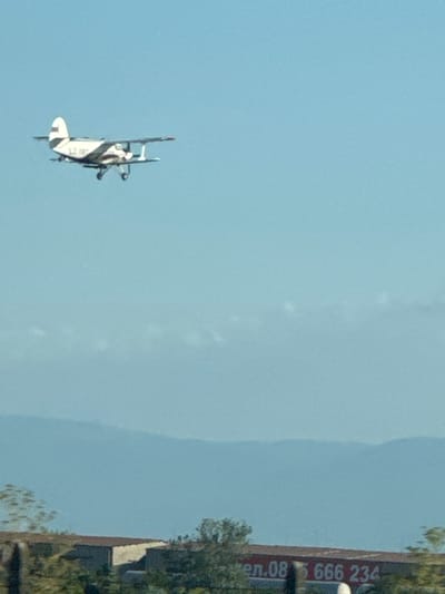 An agricultural biplane, possibly an Antonov An-2, is captured in mid-flight against a clear blue sky. The aircraft is predominantly white with dark stripes and exhibits the characteristic tail fin and high-wing design of its type. Below the aircraft, in the lower portion of the frame, a long, low building with a reddish-brown roof is visible. On the side of this building, a large sign with white text is present, reading "gen.08 5 666 234". The background features a hazy, layered mountain range. The scene appears to be an outdoor setting, potentially in or near the city of Benkovski, Bulgaria, given the presence of the building and the landscape.