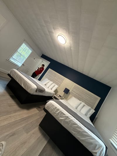 The image depicts a bedroom in a structure potentially located in West Ocean City, United States of America. The room contains two beds with white linens and grey throws, set against a blue accent wall with a wooden headboard. A person in a red shirt and dark pants stands near a partially open door, appearing to be looking at a device in their hands. A window with blinds is visible in the background, providing natural light. The room's interior includes light-colored wood-look flooring and a textured white ceiling with a central light fixture.