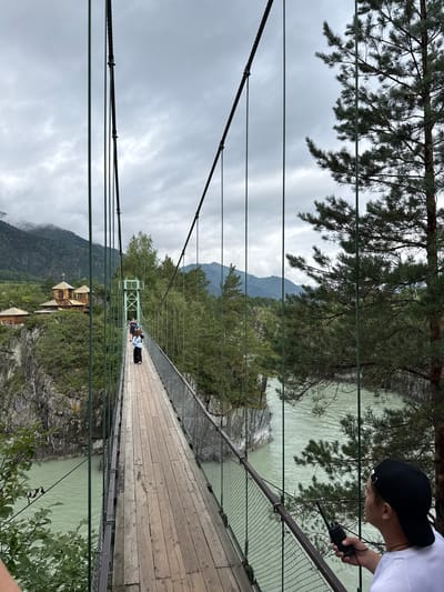 A group of people are walking across a suspension bridge spanning a jade-colored river in Chemal, Russia. The bridge has a wooden deck and metal railings. On the left side of the river, there is a cliff with a wooden building complex featuring a church with a golden dome. Lush green trees and mountains form the backdrop under a cloudy sky. In the foreground on the right side of the bridge, a person wearing a black baseball cap and a white t-shirt holds a black radio.