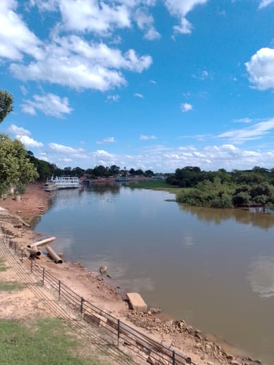 The image shows a wide, calm river in Cáceres, Brazil, under a bright blue sky with scattered white clouds. On the left bank, a large white boat is docked. The shore is made of reddish-brown earth and has a metal fence running along it. Several large pipes are lying on the bank near the water. Trees and lush green vegetation line the opposite bank, with more buildings and trees visible in the distance. The water reflects the sky and the surrounding greenery.