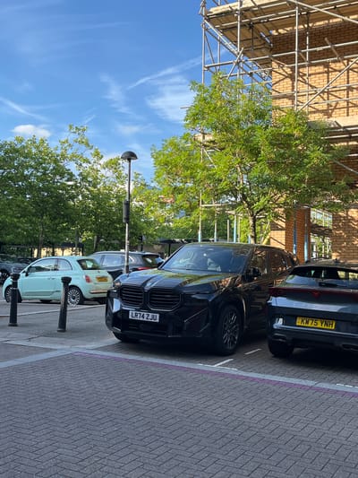 An outdoor, eye-level, medium shot captures a street scene in Milton Keynes, United Kingdom. The foreground is dominated by a dark-colored BMW SUV parked on a herringbone brick paved area, with a bright yellow UK license plate reading "LR74 ZJU". To the right of the BMW, a dark grey Cupra SUV is also parked, its rear visible with a yellow UK license plate reading "KW75 YNH" and the "CUPRA" logo on the tailgate.
In the mid-ground, a light blue Fiat 500 is parked behind the BMW, with a yellow license plate. Adjacent to it, a dark-colored SUV is parked. A modern black lamppost stands between the BMW and the Fiat. Lush green trees with dense foliage line the street, with some branches obscuring parts of the background.
The background features a brick building under construction, covered in scaffolding. The sky above is a clear blue with some wispy white clouds. The overall impression is a contemporary urban setting with parked vehicles and ongoing construction.