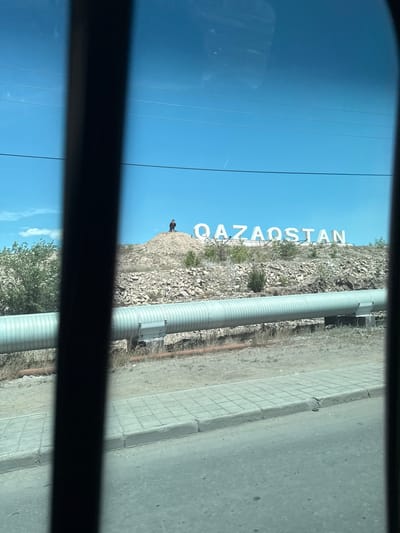 This is a medium-shot, eye-level, outdoor photograph taken through a window with vertical bars obscuring parts of the view. The image captures a wide, panoramic scene with a clear blue sky above, dotted with a few wispy white clouds. In the middle ground, on a rocky, arid hillside, stands a large white sign spelling out "QAZAQSTAN" in capital letters. To the left of the sign, on top of the hill, a dark, silhouetted figure stands on a small mound.
In the foreground, a large, light-colored industrial pipe runs horizontally across the frame, supported by metal structures. Below the pipe, there is a paved area with square tiles, suggesting a sidewalk, bordered by a curb. Beyond the sidewalk is a grey asphalt road. The overall environment appears dry and sparsely vegetated, consistent with a steppe or semi-desert landscape.
The location is identified as Jezqazğan, Kazakhstan, and the scene likely represents an entrance or a landmark for the city. The photograph was taken during daylight hours, indicated by the bright sky and shadows.