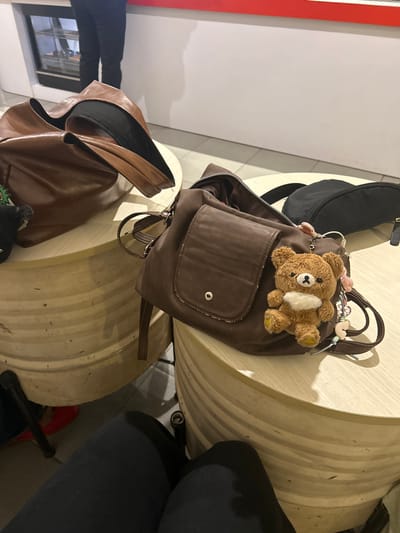 In Makassar, Indonesia, two brown leather handbags are placed on circular, wooden tables. The handbag on the right is a smaller crossbody bag, adorned with a fluffy brown bear keychain and a few pink flower charms. It rests next to a black zippered pouch. The larger handbag on the left is a tote bag, partially open, revealing a black interior. The tables are positioned in what appears to be an indoor waiting area, possibly a cafe or lounge, with a person's legs visible in the foreground, clad in dark pants. A glimpse of a person's legs and black pants is also seen in the background, standing near a wall with a red stripe. The floor is tiled, and the lighting suggests an indoor setting during daylight hours.