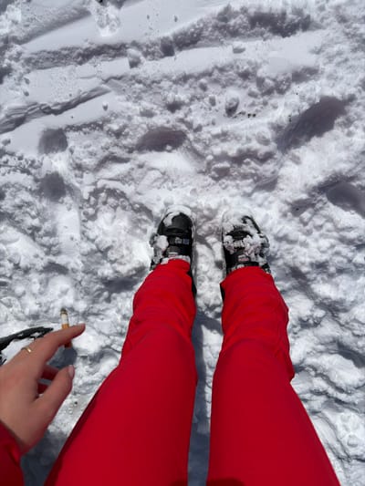 An overhead view captures a person's lower extremities and left hand in a snow-covered environment within La Clusaz, France. The individual wears bright red ski pants and black alpine ski boots, the tops of which are covered with snow. The left hand, featuring a thin gold ring on the ring finger and light-colored nail polish, holds a lit cigarette with a visible ash column between the index and middle fingers. The surrounding snow exhibits varied textures and shallow indentations, consistent with disturbed ground.