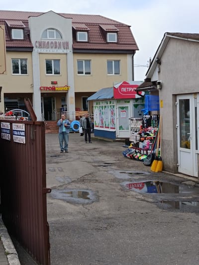 The image captures an outdoor scene in front of a commercial building, likely a small shopping center or market, with Cyrillic signage indicating it is located in the village of Smilavichi within the Smilavichy rural council of Belarus.
In the foreground, a rusty brown corrugated metal gate is partially visible on the left. The ground is paved with asphalt and shows signs of recent rain, with several puddles reflecting the surrounding environment. A woman in a light blue denim jacket and jeans stands in the middle of the paved area, holding what appears to be a smartphone to her ear. Behind her, a man in a dark jacket and grey pants stands near a display of household goods.
The building in the background has a beige facade with a prominent white section above the entrance. Cyrillic text on the building reads "СМІЛОВИЧИ" (Smilavichi) and "ТОРГОВЫЙ ДОМ" (Trading House). Below this, a red and yellow sign reads "СТРОЙМАРКЕТ" (StroyMarket), suggesting a construction and building materials store. To the right of the main building, there is a smaller kiosk or stall with a red awning that says "ПЕТРУШКА" (Petrushka). This stall displays various household items, including brooms, mops, gardening tools, and other small goods. A blue tarp is draped over the stall. The sky is overcast.