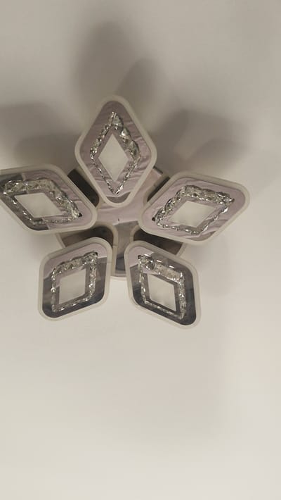 The image shows a modern, star-shaped chandelier with multiple diamond-shaped light fixtures, each containing sparkling crystal accents. The chandelier is mounted on a white ceiling, and a soft shadow is cast onto the ceiling from the fixture. The lighting is subdued, suggesting it may be evening or that the light is not fully on. The image provides a close-up view of the chandelier, with the majority of the frame dedicated to the fixture and the ceiling. There are no people or other objects visible in the image, and the background is a plain white ceiling. The location is identified as the city of Criuleni, Moldova.