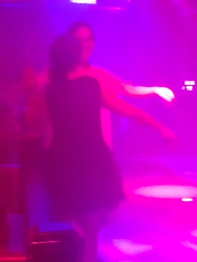 The image shows two individuals in a dimly lit setting, illuminated by vibrant pink and purple lights, suggesting a nighttime or indoor event like a club or party. One person in the foreground is wearing a dark, possibly black, dress that flares out at the bottom. Their posture suggests they are dancing or posing. Another person is partially visible behind and to the right of the first person, their features obscured by the lighting and their pose. The background is a blur of magenta and violet, with some bright white highlights that could be from stage lights or other illumination. The overall atmosphere appears energetic and festive. There is no visible text in the image. The location context is Nanning, China.