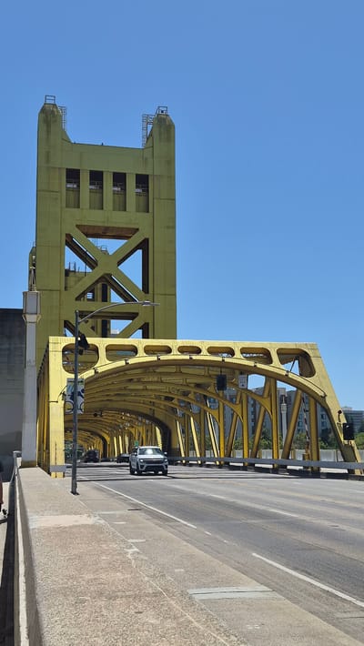The image depicts the Tower Bridge in West Sacramento, California, United States of America. The bridge is characterized by its distinctive yellow, drawbridge structure with two prominent towers. In the foreground, a roadway is visible with two lanes of traffic. A white SUV is driving on the road, and other vehicles are in the distance. A pedestrian walkway is present on the left side of the road. The sky is clear blue, and the sun is shining, casting shadows on the bridge and road. The bridge's intricate metalwork is clearly visible, as are the surrounding buildings and trees.