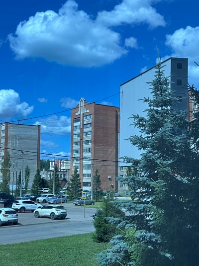 This image depicts an outdoor scene in the city of Mariinsky, Russia, likely a residential area on a clear day with a bright blue sky and scattered white clouds. In the foreground, a large, mature evergreen tree with bluish-green needles partially obstructs the view. Behind the tree, a parking lot is filled with various passenger vehicles.
Dominating the background are several multi-story apartment buildings. To the left, a tall, rectangular building with a light-colored facade and numerous windows stands prominently. Adjacent to it, to the right, is another tall apartment building constructed of reddish-brown brick, also featuring many windows and a decorative gable-like structure at its peak. Further in the background, more apartment buildings of varying heights and designs are visible, along with trees and streetlights. A pedestrian crossing sign is visible on a pole in the parking lot. The overall impression is one of a typical urban residential landscape.
