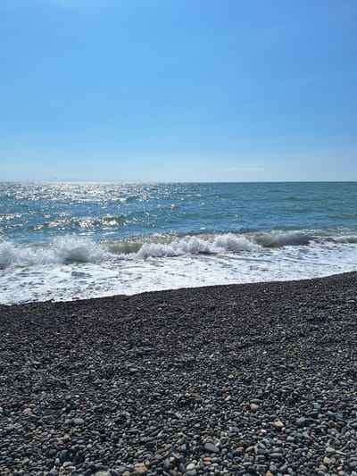 The image depicts a pebbled beach on a sunny day in Kobuleti, Georgia. The foreground is filled with a dense field of dark grey and multi-colored pebbles, extending from the bottom of the frame upwards. White foam from gentle waves laps at the edge of the pebble shore. The middle ground features the expanse of the sea, with the surface shimmering under the bright sun, creating a dazzling reflection. Waves are breaking, with whitecaps and churning water visible. The horizon line is distinct where the sea meets the clear, bright blue sky. The sky is mostly cloudless, with a hint of lighter haze near the horizon. There are no people or man-made structures visible in the frame. The scene captures a tranquil natural environment at the coast.
