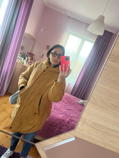 A woman with glasses takes a mirror selfie. She is wearing a tan-colored parka and blue jeans. A pink rug and purple curtains are reflected in the mirror. A white lamp hangs from the ceiling, and a window lets in natural light. Various items are on a shelf or dresser behind her. She holds a red phone, presumably used to take the photo.
