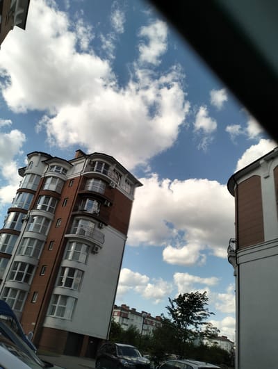 The image shows a view of buildings in Krykhivtsi, Ukraine, under a blue sky with fluffy white clouds. A multi-story building with a reddish-brown and white facade is on the left. It features multiple curved balconies with black railings. To the right, part of another building with similar architectural elements is visible. In the foreground, there are cars parked and a dark object is partially obstructing the view. The background shows trees and other residential buildings. A flock of birds is visible in the sky.