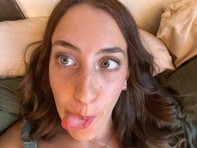 An overhead, inverted shot captures a young person, likely female, lying down, possibly in a bed, in Melbourne, Australia. The subject is seen from the chest up, with their head positioned upside down in the frame. They are making a playful expression, sticking out their tongue and crossing their light-colored (hazel or green) eyes, which are wide open.

Their long, wavy, dark brown hair spreads out around their head against a lighter-colored pillow and a darker green sheet or blanket. A small silver stud piercing is visible on their nose, and a small hoop earring can be seen in their right ear. Part of a green, possibly lace, garment strap is visible near their shoulder. The lighting suggests an indoor setting, and the overall mood is lighthearted and humorous due to the exaggerated facial expression. No visible text is present in the image.