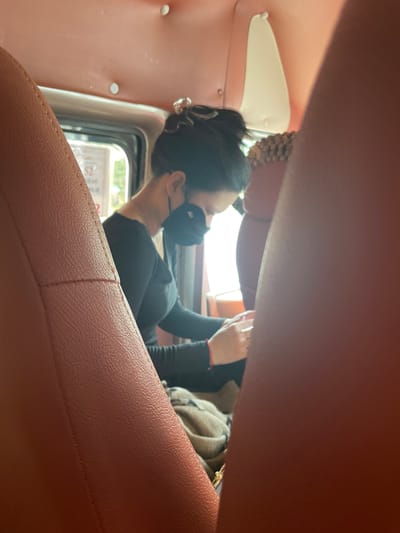 The image captures an interior view of a vehicle, likely a bus or van, seen from the perspective of a passenger seated behind another individual. The dominant colors are warm tones of red and pink, seen in the upholstery of the seats and the ceiling. In the mid-ground, a person wearing a black long-sleeved shirt and a black face mask is seated, looking down at their hands which are holding a mobile device. Their hair is styled in a bun with a clear clip. A red bracelet is visible on their left wrist. A patterned blanket is draped over their lap. Outside the vehicle, through a window, greenery and some text on a sign are partially visible, suggesting an urban or semi-urban setting. The scene appears to be candid, with the focus on the person using their phone while traveling. The location is identified as Ben Cau, Vietnam.