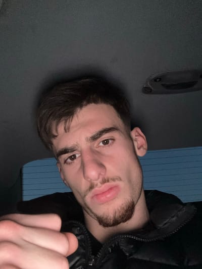 A low-light portrait captures a young man, likely in his late teens or early twenties, positioned within what appears to be the interior of a vehicle in Souffelweyersheim, France. His head is tilted to the left, and he looks directly at the camera with a neutral to slightly pouting expression, characterized by slightly parted, full lips. He has short dark hair, a neatly trimmed goatee, and brown eyes. He is wearing a dark, possibly black, puffer jacket.
The background primarily shows the dark grey ceiling or headliner of the vehicle, with an overhead console featuring a light or control panel. Clearly visible on this console is the text "B1". Behind his head, to the right of the frame, are vertical blue elements, possibly blinds visible through a window or a textured wall section. His right hand, partially clenched, is visible in the bottom right corner of the frame. The dim lighting conditions suggest an indoor setting or nighttime, with his face well-lit by an apparent flash or direct light source.