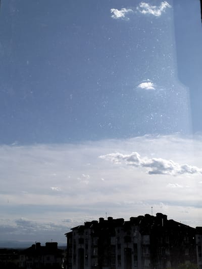 The image shows a view from inside a building, looking out over a partially overcast sky and a section of Krykhivtsi, Ukraine. The sky transitions from a clear blue at the top to a more cloudy, overcast lower portion. A few small, puffy clouds are visible. The view includes the silhouette of a building complex, possibly residential, with multiple sections and varying heights. Several antennae are visible on the roof of the building. The lower portion of the image, outside the window, shows more buildings and a hint of distant terrain. The window pane shows signs of wear and smudges.