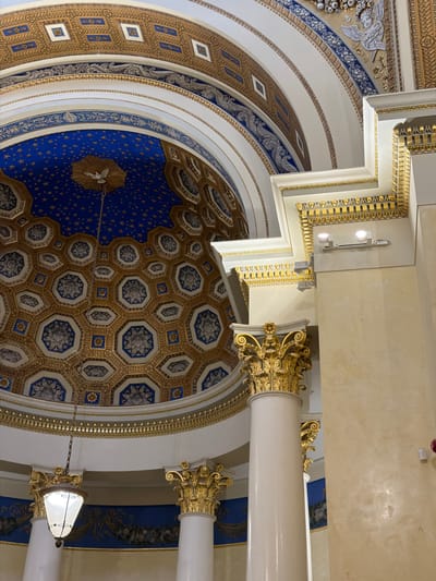 The image shows an interior view of a grand, ornate ceiling and columns. The ceiling is a dome, decorated with intricate patterns in gold and blue. The dome features a starry sky motif with a dove in the center. Surrounding the dome are bands of decorative friezes, some with square motifs and others with floral or foliate designs.
The columns are tall and cylindrical, made of a smooth, light-colored material, likely marble or plaster. They are topped with elaborate gold Corinthian capitals, featuring scrolling acanthus leaves. Decorative gold moldings and cornices are visible throughout the scene, accentuating the architectural elements. A hanging lantern with a glass shade is suspended from the ceiling by chains.
The overall impression is one of opulence and classical architecture. While the provided metadata suggests a location in "посёлок дачного хозяйства «Архангельское», Russia," there are no specific visual cues within the image itself that confirm this exact location. The scene is devoid of people and any discernible actions or interactions. The lighting appears to be artificial, likely from the overhead lamp and possibly other concealed sources, creating a warm and ambient atmosphere.