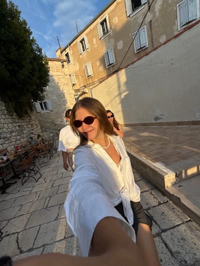 In the city of Split, Croatia, a selfie is taken on a cobblestone street lined with stone buildings and outdoor seating for a restaurant. The woman in the foreground, wearing sunglasses and a white shirt, is smiling at the camera. Behind her, two other individuals are walking away from the camera. The sun is setting, casting long shadows across the street and buildings, and the sky is a clear blue with a few wispy clouds. The scene appears to be part of an old town area, with the architecture suggesting a historical setting.