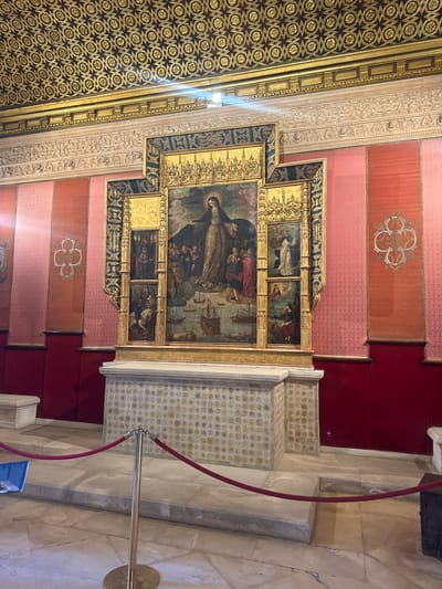 The image depicts a richly decorated interior space, likely within a historical building in Seville, Spain. Dominating the scene is a large, ornate altarpiece featuring a central painting of a woman, possibly a religious figure, surrounded by a crowd, with ships visible in the background. The altarpiece is framed by gold and dark green decorative elements, with smaller religious scenes depicted in vertical panels on either side of the central painting.
The altarpiece rests on a decorative tiled pedestal, which is itself situated on a stone platform. The walls surrounding the altarpiece are covered in a pink textured wallpaper with a repeating geometric pattern, interspersed with vertical panels of a deeper red fabric adorned with stylized floral motifs. Above the walls, a highly decorative golden ceiling with a repeating geometric pattern is visible.
In the foreground, a red velvet rope is strung between brass stanchions, cordoning off the area in front of the altarpiece, suggesting it is a protected exhibit or sacred site. The floor is made of marble tiles. The overall impression is one of opulence and historical significance, characteristic of many religious and palatial buildings in Seville.