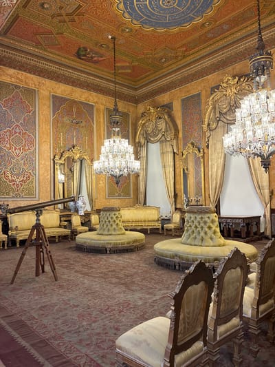 The image depicts a lavishly decorated room, likely part of a palace or historic building, possibly located in Üsküdar, Turkey. The room is adorned with ornate gold accents, intricate ceiling frescoes with celestial motifs, and detailed patterned wallpaper. Dominating the scene are plush, yellow upholstered furniture, including several curved sofas and round, tufted ottomans. Two large crystal chandeliers hang from the ceiling, illuminating the opulent surroundings. A vintage telescope on a tripod stands in the foreground, suggesting a place of study or contemplation. Mirrors with elaborate gold frames are strategically placed on the walls. The floor is covered by a large, patterned rug with muted colors. The overall impression is one of grandeur and historical significance.