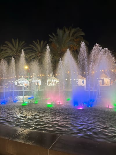 The image depicts a nighttime scene at a fountain. The fountain is illuminated with vibrant colored lights, including blue, pink, and green, which reflect off the water. Several palm trees are visible in the foreground, their fronds creating a textured silhouette against the dark background. In the background, there are structures that appear to be tents or stalls, possibly part of a market or event, also lit with warm, yellow lights. People are visible inside or near these structures, suggesting an active gathering. The water from the fountain is spraying upwards, creating a dynamic and refreshing visual effect. The overall atmosphere is festive and lively, indicating an evening event in a pleasant, likely warm, climate. The presence of palm trees and the overall ambiance are consistent with a Mediterranean location like Sant Antoni De Portmany, Spain. No visible text is present in the image.