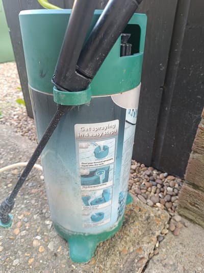 A vertical, slightly angled medium shot shows a green and white plastic pressure sprayer standing on a concrete patio next to a dark wooden fence and a gravel path. The sprayer is cylindrical with a green base and cap. A black spraying wand with a green nozzle extends from the side, resting against the body of the sprayer. A label on the sprayer provides instructions with small illustrations. The label reads "Get spraying in 4 easy steps" and details steps for pouring liquid, selecting a spray setting, and pumping the sprayer. The environment suggests an outdoor setting, possibly a garden or yard in Huntingdonshire, United Kingdom, given the presence of typical garden elements like a fence, gravel, and concrete. The lighting indicates daylight, casting soft shadows. There are no people or animals visible in the frame.