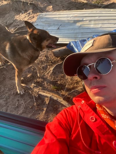 A ground-level, slightly rotated image shows a person and a dog in an outdoor setting, potentially in a rural or semi-rural area. The person is wearing a brown baseball cap, round sunglasses, and a red hooded jacket with a paisley bandana underneath. The jacket is buttoned at the top. The person is partially reclined. Next to the person stands a medium-sized, short-haired dog with light brown fur. The dog's mouth is open, showing its teeth and tongue. The ground is covered in dirt and dry vegetation. In the background, there is corrugated metal siding and parts of a blue and green striped object. The overall lighting suggests it is daytime. Based on the prompt specifying Vyritsa, Russia, the scene is most likely located in an outdoor area near Vyritsa.