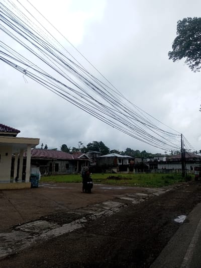 A street scene under a cloudy sky shows a person on a motorcycle centered in the frame. Dilapidated houses line the background, separated by overgrown grass. Overhead, a dense network of electrical wires spans the sky. The road is cracked and uneven, with a dark, coarse texture in the foreground. A building sits to the left of the frame, while a tree is visible on the upper right.