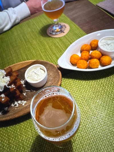This is a close-up, overhead shot of a table setting with food and drinks. In the foreground, a glass of amber-colored beer with a foamy head sits on a green textured placemat. To the left, a wooden platter holds a pile of dark, bite-sized croutons sprinkled with white grated cheese, next to a small white ramekin filled with a creamy white sauce. In the background, a white serving dish contains several round, golden-orange fried appetizers and a small ramekin of white sauce. Another glass of beer is visible in the upper background, placed on a coaster. A person's arm, wearing a white long-sleeved shirt, is visible on the far left, suggesting a dining or social setting. The overall impression is of a casual meal or appetizer spread. The location is not specified in the image.
