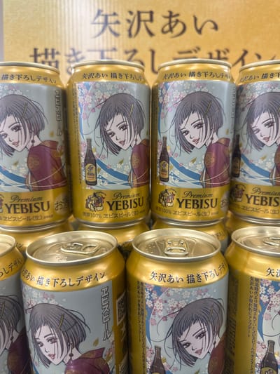 The image shows a stack of gold-colored beer cans with a collaborative design featuring artwork by manga artist Ai Yazawa. Each can features an anime-style female character dressed in a kimono against a background of flowers. The can also has the word "YEBISU" prominently displayed, indicating the brand of beer. Japanese text is visible on the cans. There's a blurry background with Japanese text, giving the impression that the cans might be displayed or photographed on a promotional material or table. There's no indication in the image regarding the city of Nakano, Japan.