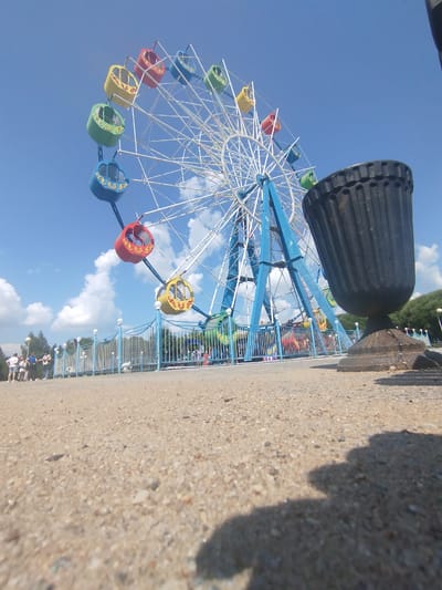 This image depicts a ferris wheel in an amusement park, likely located in Yalutorovsk, Russia, given the context. The ferris wheel is a prominent structure with blue supports and a white frame, featuring brightly colored gondolas in red, yellow, and green. In the foreground, a dark, ornate trash receptacle stands to the right, casting a shadow on the sandy ground. Behind the ferris wheel, a blue and white fence encloses the amusement park area, with several people visible walking and standing near the entrance. Trees line the background, suggesting a park or outdoor recreational setting. The sky is a clear blue with scattered white clouds. The perspective is low, emphasizing the scale of the ferris wheel and the ground texture.