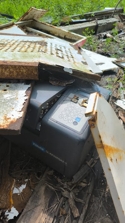 A Konica CL-KP32EQA2-3 time clock is discarded amidst debris in Turgoyak, Russia. The time clock, a dark gray electronic device with a keypad and a small screen, is partially covered by rusted metal sheets and decaying wooden planks. The surrounding environment appears to be an outdoor area with overgrowth of green vegetation, suggesting it is either a junkyard or an abandoned location. The scene is littered with remnants of what appear to be industrial equipment or building materials, indicating a state of disuse and neglect.