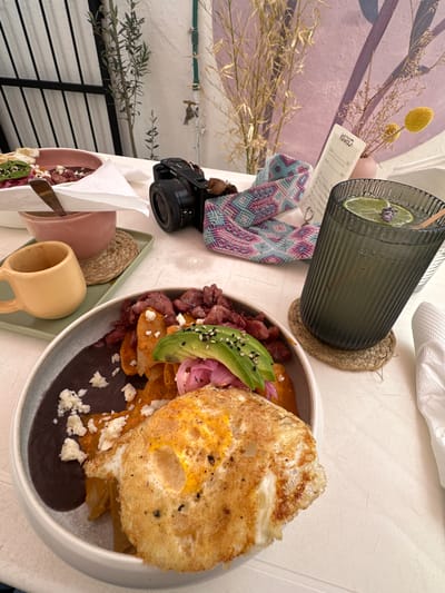 A close-up, overhead shot captures a table setting at a restaurant in Zacatecas City, Mexico. The scene is dominated by a plate of chilaquiles topped with a fried egg, avocado, and pickled red onions. Beside the chilaquiles is a bowl of black beans with crumbled white cheese. A small yellow mug sits on a green tray to the left, next to a pink bowl. In the background, a camera rests on a patterned textile, with a glass of iced beverage featuring lime slices and a lavender sprig. A menu, a vase with dried plants and flowers, and a wall with abstract art complete the composition. The table is covered with a white tablecloth, and a black metal fence is visible in the upper left corner, suggesting an outdoor or semi-outdoor dining area.