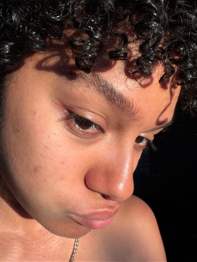 A close-up, side-profile shot of a person's face and upper body is captured in natural sunlight. The subject has dark, curly hair that is wet and shiny, with prominent curls framing their forehead. The sunlight casts a shadow of the hair across their forehead and cheek. The person's skin appears to be of a darker complexion with some subtle blemishes and pores visible, particularly on the cheek. Their eyes are dark brown and appear slightly closed or looking downwards. The bridge of their nose and lips are also visible, with the lips appearing slightly parted. A thin, gold chain necklace is visible around their neck. The background is completely black, indicating a dark, possibly indoor or shadowed environment. The image does not contain any information that would allow for a determination of its location in the city of Champs-sur-Marne, France.
