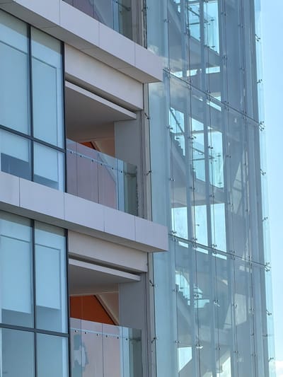 The image displays a detailed, upward-angled view of a modern multi-story building facade in Fraccionamiento Los Heroes Queretaro, Mexico, under clear daylight conditions. The left portion of the building features light-colored, possibly off-white or light gray, horizontal paneling delineating floor levels. These panels frame large, rectangular windows with dark-colored mullions, reflecting a light blue sky. Below one of the upper panels, a recessed balcony area is visible, enclosed by a clear glass balustrade secured with small metal fixtures. An interior wall within this balcony appears to be an orange-brown color.
The right portion of the image is dominated by a transparent, multi-story glass curtain wall, characteristic of a point-fixed glazing system, with numerous circular spider fittings securing the individual glass panels. Behind this transparent facade, the internal structure of a staircase is discernible, composed of light-colored, possibly concrete or metal, risers and stringers, ascending diagonally. Light filters through and reflects off the glass surfaces, creating varied tones and shadows within the stairwell. No individuals or specific actions are visible within the frame.