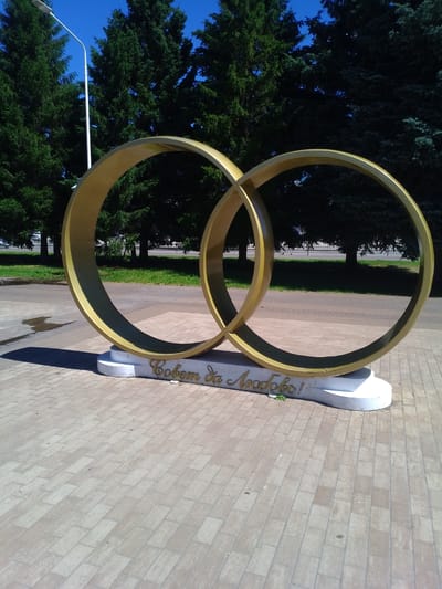 The image shows a monument in the city of Otradny, Russia, featuring two interlocking golden rings on a white concrete base. The monument is situated on a paved area, possibly a walkway or plaza, made of light-colored rectangular bricks. Behind the monument, there is a row of tall evergreen trees. In the background, a street is visible with a sidewalk and some buildings. A modern white lamppost stands to the left of the monument. The sky is clear and blue. The inscription on the base of the monument appears to be in Cyrillic script. The lighting suggests it is a sunny day, casting shadows on the ground.