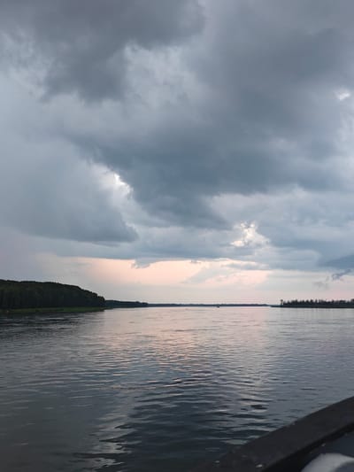The image depicts a wide river with choppy water reflecting a dramatic, overcast sky. Dark, heavy clouds dominate the upper portion of the frame, with some lighter patches breaking through towards the horizon. A dense, dark green forest lines the left bank of the river, stretching into the distance. The right bank is less distinct, appearing as a thin line of trees in the background. The water's surface is rippled, indicating some wind or current. The scene appears to be a natural landscape, possibly along the Angara River near Kazachinskoe, Russia, given the typical geography of the region. No people or specific man-made objects are prominently visible, except for a dark, blurred railing in the foreground, suggesting the photo was taken from a boat or pier.