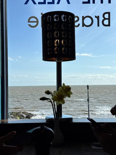 The image depicts an interior view from a restaurant or brasserie identified by the reversed window text "THE VEEX Brassiere" (likely "The View Brasserie"), overlooking the North Sea in Felixstowe, United Kingdom.
In the immediate foreground, a dark, textured table surface is visible. Two sets of silhouetted hands suggest the presence of at least two individuals. On the left, a hand holds a dark, rectangular object, while on the right, a hand grips a smartphone.
Central to the foreground is a dark, cylindrical table lamp with a chrome-colored stem and base. Its shade features a patterned cutout design, through which the illuminated bulb casts a warm light. To the right of the lamp, a small white vase contains a bouquet of yellow daffodils, comprising three open blooms and two closed buds. A dark, ornate object, possibly a condiment shaker, is partially visible on the table to the left.
Beyond the window, a light blue sky with scattered white clouds transitions to the expansive, choppy, grey-brown waters of the North Sea. The horizon line is clearly visible. To the left, a rocky breakwater extends into the water. In the mid-distance, several small, red/orange buoys are dispersed across the water's surface, and a thin, vertical pole structure, resembling a distant lamp post or pier element, rises from the sea towards the right. The exterior natural light is strong, backlighting and silhouetting the interior foreground elements.