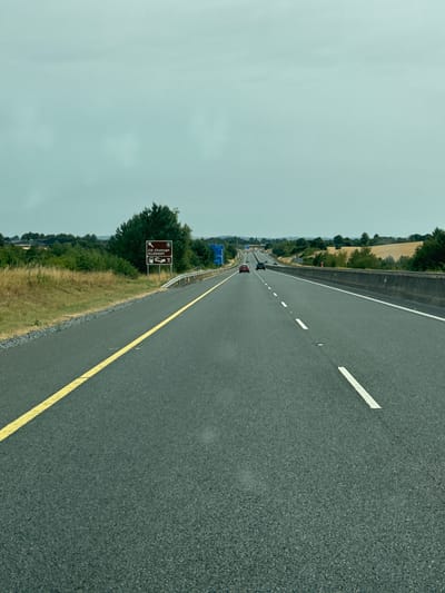 The image shows a view from a vehicle driving on a multi-lane highway. The road is asphalt with yellow and white lane markings. The sky is overcast and a pale blue-green. On the left side of the road, there is a signpost indicating "Cill Chainnigh KILKENNY" with directional arrows and icons for fuel, lodging, and a restaurant. Beyond the road, there are fields with dry grass and scattered trees. A few cars are visible on the highway in the distance. The location is identified as a highway near Glaunta, Ireland.