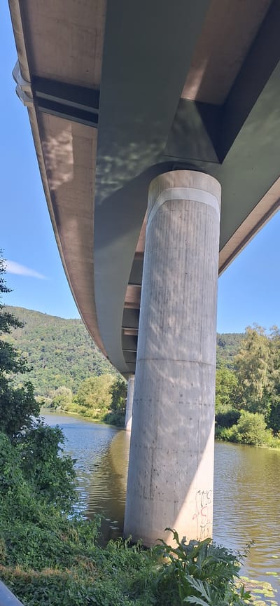 The image shows a concrete pillar supporting a bridge over a river in Fachbach, Germany. The pillar is cylindrical and textured with vertical lines. Two light-colored bands encircle the top of the pillar. Graffiti in red and black is visible on the lower part of the pillar. The river flows gently, with reflections of the bridge and trees on its surface. Lush green trees and vegetation cover the hilly landscape in the background. The sky is clear and blue.