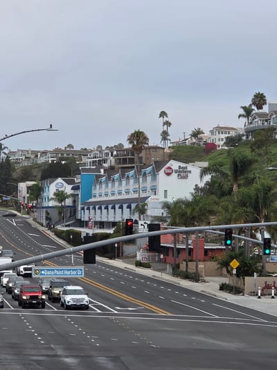 The image shows a street scene in Dana Point, California, USA, on an overcast day. The foreground features a multi-lane road with several cars stopped at a traffic light. A blue and white sign indicating "Dana Point Harbor Dr" is visible. Beyond the road, a hotel with blue and white architecture is situated on a hillside. The hotel has a sign that reads "Best Western PLUS". In the background, several houses and buildings are scattered across the hilly landscape, with palm trees dotting the scene. Traffic lights are visible, with some showing green. A speed limit sign indicating 35 is also present.