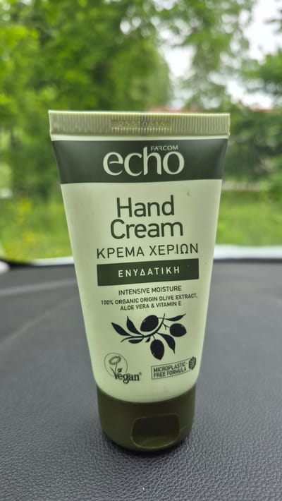A light green tube of Echo Hand Cream with a dark green cap is the primary subject of the image. The tube has black and white text that reads "echo," "FARCOM," "Hand Cream," "ΚΡΕΜΑ ΧΕΡΙΩΝ," and "ΕΝΥΔΑΤΙΚΗ." Below this, it states "INTENSIVE MOISTURE 100% ORGANIC ORIGIN OLIVE EXTRACT, ALOE VERA & VITAMIN E." There is a graphic of an olive branch with olives. To the left of the olive branch graphic, there is a small circular logo that says "Vegan" with a plant image. To the right of the olive branch graphic, there is a rectangular icon that reads "MICROPLASTIC - FREE FORMULA." A batch code "25310315" is visible at the top of the tube. The tube is positioned on a dark gray textured surface, possibly a dashboard or a car interior. In the background, out of focus, is lush green foliage, suggesting an outdoor setting. The image does not provide any specific information about the city of Dagda, Latvia.