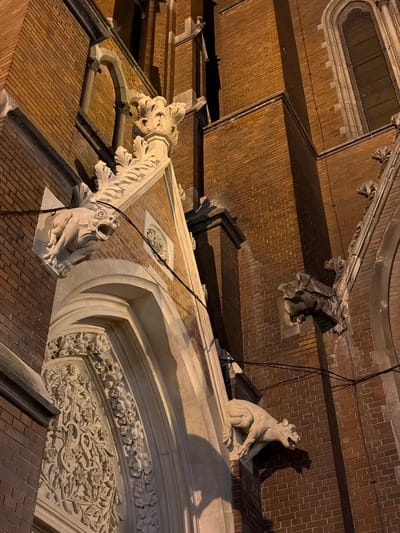 The image shows a close-up view of architectural details on a brick building, likely taken at night due to the warm, artificial lighting. The building features ornate stone carvings, including gargoyles perched on the facade. One gargoyle appears to be in a seated position with its mouth open. The brickwork is visible in various sections, with some areas in shadow. The stone details include floral patterns and decorative moldings. Based on the architecture, which includes brickwork, stone carvings, and gargoyles, it is likely taken at the Co-cathedral of St. Peter and St. Paul in Osijek, Croatia.