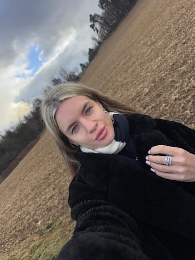 An adult female is captured in a selfie-style, tilted outdoor photograph, situated in a rural area near Regenstauf, Germany. She has long, blonde hair, blue eyes, and full lips, and is looking directly at the camera. She wears a dark, possibly faux fur, coat and a white and navy striped scarf. Small stud earrings are visible in her ears. Her left hand (viewer's right) is prominent, displaying light pink nail polish and a large, multi-band ring adorned with numerous stones on her ring finger. The background consists of a vast, brown, tilled field, leading to a distant treeline composed of both leafless deciduous trees and dark evergreen trees. The sky above is largely overcast with grey clouds, though patches of blue are visible. The overall scene suggests a cool, possibly late autumn or winter day.