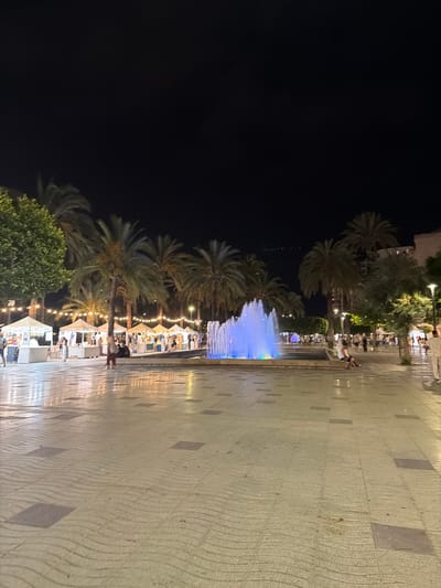 The image depicts a nighttime scene in the city of Sant Antoni De Portmany, Spain, likely on a promenade or public square. A vibrant fountain with blue and purple lighting is the central focus, surrounded by a paved area. Palm trees and lush green trees line the background, with several market stalls or vendor tents set up along the left side, illuminated by string lights. People are visible walking, browsing the stalls, and sitting in the vicinity of the fountain. The ground is paved with large square tiles, some of which have a wave-like pattern. Buildings and streetlights can be seen in the distance.
