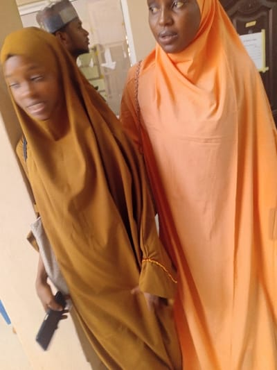 The image displays an indoor scene featuring three individuals. In the foreground, two females stand side-by-side. The female on the left, wearing a brown full-length abaya and matching hijab, holds a black smartphone in her right hand and has a light-colored strap over her left shoulder. Her face is slightly blurred, with her mouth partially open. The female on the right, wearing an orange full-length abaya and matching hijab, looks slightly upwards and to her left, with her mouth also partially open. A light-colored strap is visible on her left shoulder. In the blurred background, a male wearing a patterned cap and a light-colored shirt is visible in profile, looking to the right. The environment consists of light-colored walls, with a dark-framed doorway and a white sheet of paper or notice affixed to it visible in the background. The specific location within Kwazailewa, Nigeria, cannot be determined from the visual information presented.