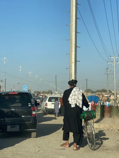 The image depicts a street scene in Arghandab, Afghanistan. A man wearing traditional Afghan clothing and a turban stands on a dirt shoulder beside a busy road. He is holding a bicycle with a green container attached to it. A white towel is draped over his shoulder. In the background, several cars are lined up in traffic. There are also metal containers and other debris on the side of the road. Tall concrete electricity poles are visible in the background. The sky is clear and blue.
